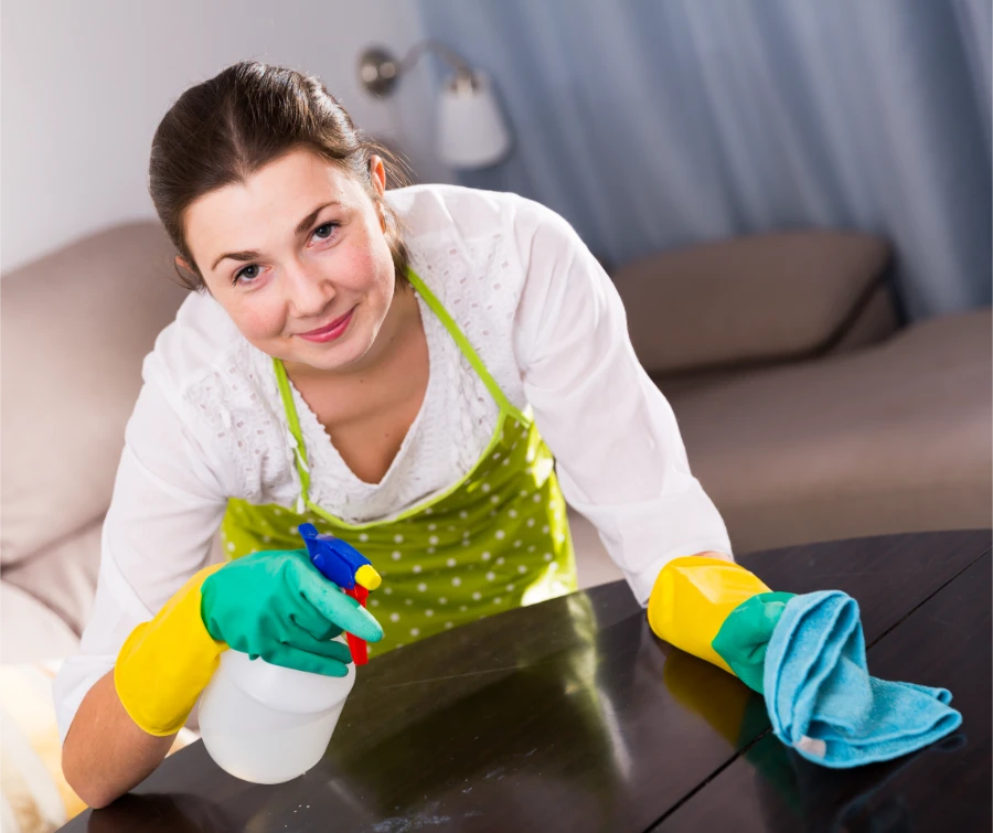 Professional deep cleaning service woman smiling while spraying and wiping
dark table surface with microfiber cloth and rubber gloves in living room